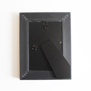 May include: A rectangular black picture frame shown from the back, featuring a black backing board, two metal hanging hooks, and a hinged support stand. The frame border is decorated with small, white, hand-drawn arrow-like patterns at corners.