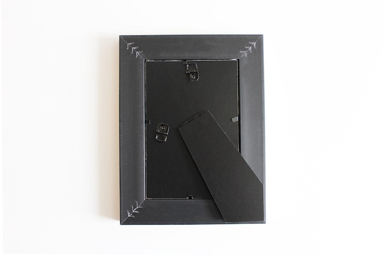 May include: A rectangular black picture frame shown from the back, featuring a black backing board, two metal hanging hooks, and a hinged support stand. The frame border is decorated with small, white, hand-drawn arrow-like patterns at corners.