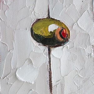 May include: An oil painting on canvas featuring a single green olive with a red pimento center, skewered on a thin vertical stick. The background consists of thick, textured white impasto paint strokes. A signature and the year 2024 appear at the bottom.