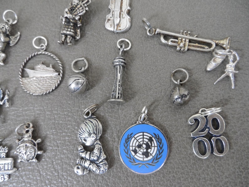 Może przedstawiać: Kolekcja srebrnych zawieszek, w tym statek, koszyk&oacute;wka, trąbka, baletnica, modlące się dziecko i emblemat ONZ. Inne zawieszki to dudziarz, skrzypce, globus i liczba 2000.