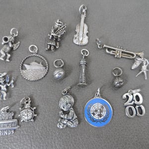 Może przedstawiać: Kolekcja srebrnych zawieszek, w tym cherubin, szkocki dudziarz, skrzypce, trąbka, statek wycieczkowy, iglica Space Needle w Seattle, piłka do koszyk&oacute;wki, baletnica, biedronka, dziecko, emblemat ONZ i liczba 2000.