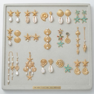 May include: A collection of gold-toned earrings featuring nautical motifs including starfish, seashells, and pearls, arranged on a gray display tray with a plaque reading THE SEA COLLECTION - FULL ARCHIVE. The set includes various drop and stud styles.