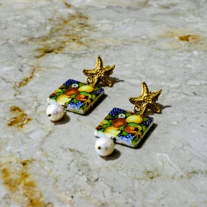 May include: A pair of gold-colored starfish earrings with square, hand-painted ceramic pendants. The pendants feature a colorful fruit design and are accented with a dangling white pearl. The earrings are set against a marble background.