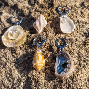 Peut inclure: Cinq boucles d'oreilles individuelles composées de coquillages naturels et de petites perles blanches. Chaque pièce est fixée à un anneau en métal argenté ou doré. La collection comprend une huître plate, un conque spiralé, une coquille Saint-Jacques, un cône jaune et une coquille d'ormeau irisée avec plusieurs perles.