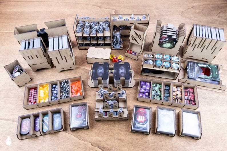 May include: A collection of laser-cut wooden organizers and storage boxes for board game components. The set includes various containers for cards, tokens, dice, and miniatures, arranged on a wooden surface. The organizers are designed to keep game pieces organized.