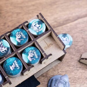 May include: A wooden game token organizer with various tokens. The tokens are teal and white with different labels, including "Drone", "Adult", and "Larva". The organizer is labeled "Queen". Dice are visible in the background.