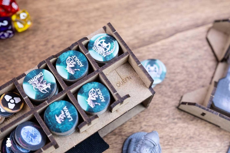 May include: A wooden game token organizer with various tokens. The tokens are teal and white with different labels, including "Drone", "Adult", and "Larva". The organizer is labeled "Queen". Dice are visible in the background.