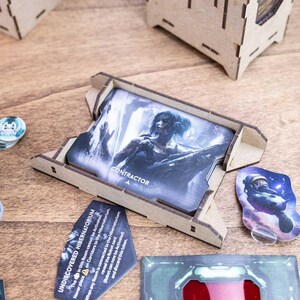 May include: A tabletop game with various components, including cards, tokens, and wooden game piece holders. The cards feature illustrations and the word "CONTRACTOR." Other components include a miniature astronaut and a card with the text "UNDISCOVERED HIBERNATORIUM."