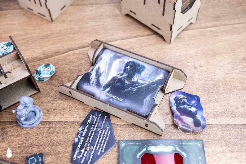 May include: A tabletop game with various components, including cards, tokens, and wooden game piece holders. The cards feature illustrations and the word "CONTRACTOR." Other components include a miniature astronaut and a card with the text "UNDISCOVERED HIBERNATORIUM."