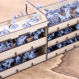 May include: A wooden organizer with multiple compartments filled with miniature figurines. The figurines are various shades of gray and are arranged in rows. The organizer has a natural wood finish and is designed for tabletop gaming.