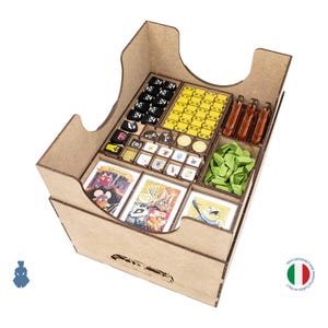 May include: A wooden board game organizer insert featuring multiple compartments. It contains various game components including black and yellow tokens, small wooden pieces, green leaf-shaped tokens, and a set of illustrated playing cards.
