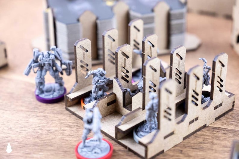 May include: Miniature gray figures and laser-cut wooden structures are arranged on a wooden surface. The figures are on colored bases, and the structures have a geometric design. The scene suggests a tabletop game or hobby.