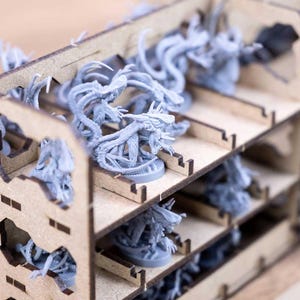 May include: A wooden organizer filled with gray miniature figures. The organizer has multiple shelves and cut-out designs. The figures appear to be fantasy creatures with tentacles and other otherworldly features. The overall theme suggests a tabletop gaming accessory.
