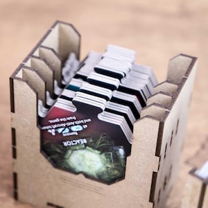 May include: Wooden game card holders filled with game cards. The holders are made of light brown wood and have a slotted design. The cards have a black and white design with some color accents and text. The text on the cards reads "REACTOR".