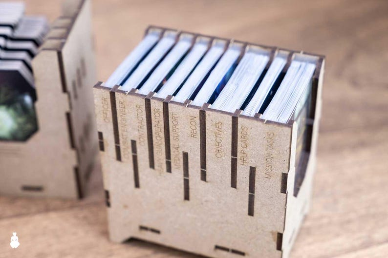 May include: Two wooden card holders with laser-cut labels. The labels include "Officer", "Combat Engineer", "Contractor", "Heavy Gun Operator", "Medical Support", "Recon", "Objectives", "Help Cards", and "Mission Tasks". The holders contain stacks of cards.