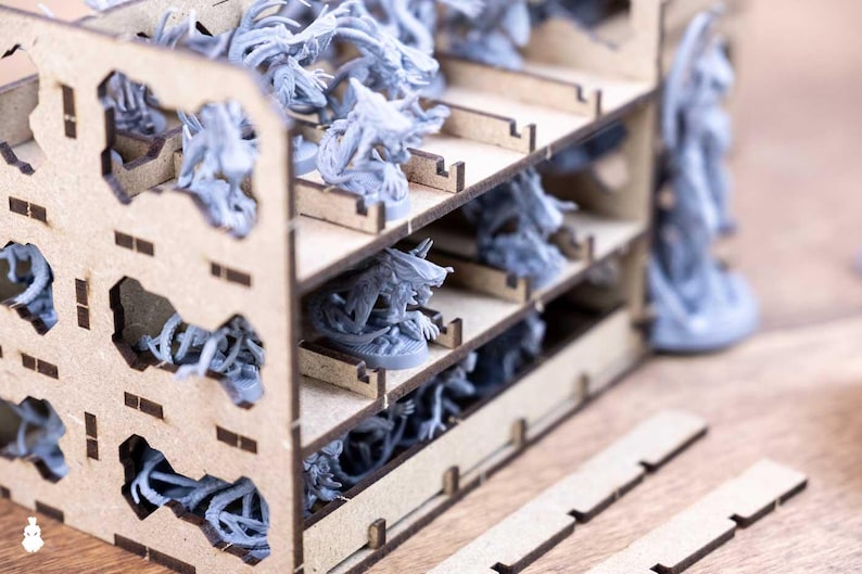 May include: A wooden organizer with multiple shelves, filled with gray miniature figurines. The organizer has cut-out shapes and is designed to hold the figurines. The figurines appear to be fantasy or sci-fi themed.