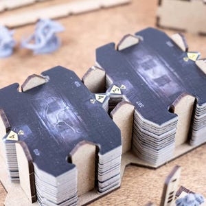 May include: Stacks of black and gray game cards with a detailed graphic design, arranged on a wooden surface. The cards have a unique shape and are part of a board game. Additional game pieces are visible in the background.
