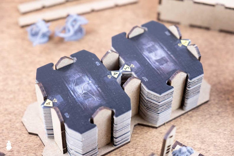 May include: Stacks of black and gray game cards with a detailed graphic design, arranged on a wooden surface. The cards have a unique shape and are part of a board game. Additional game pieces are visible in the background.