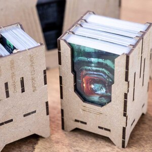 May include: Laser-cut wooden card holders with slots and text that reads "Queen Hlagh", "Robot", "Character Items", and "Support Equipment". The holders contain stacks of cards with colorful images. A miniature figure is visible.