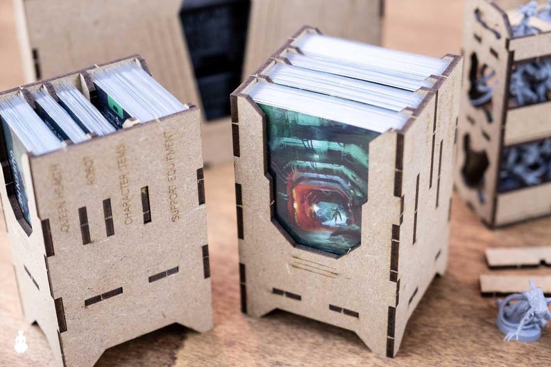 May include: Laser-cut wooden card holders with slots and text that reads "Queen Hlagh", "Robot", "Character Items", and "Support Equipment". The holders contain stacks of cards with colorful images. A miniature figure is visible.