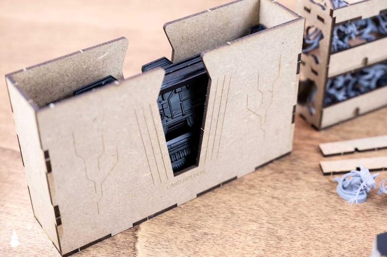May include: A set of unfinished wooden card holders, with the text "CARD HOLDERS" visible. The holders are designed with a geometric aesthetic and feature cut-out designs. Other components and miniatures are also present.