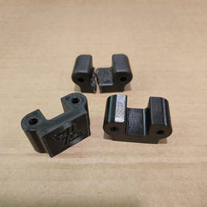Puede incluir: Tres piezas de plástico negro impresas en 3D. Dos de las piezas están conectadas entre sí. La tercera pieza tiene un pequeño logotipo en el lateral.