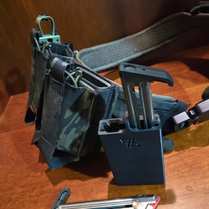 May include: A tactical belt system with camouflage pouches and a black magazine holder. A silver and red magazine is lying on a wooden surface. The belt has a green buckle and a padded strap.