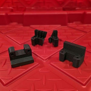 Puede incluir: Cuatro componentes de plástico negro impresos en 3D están dispuestos sobre una superficie roja. Los componentes tienen varias formas, incluyendo soportes y piezas con características de enclavamiento. Las piezas parecen ser componentes funcionales.