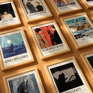 Può includere: Una collezione di poster di film incorniciati con vari titoli, tra cui "Donnie Darko", "A Farewell to Arms" e "Inception". I poster presentano illustrazioni colorate e testo, esposti in cornici di legno chiaro, disposti su una superficie di legno.