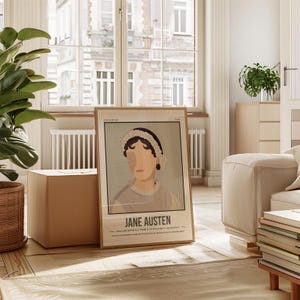 May include: A framed poster featuring a minimalist illustration of Jane Austen, with a quote from her novel "Persuasion." The poster is in a light wood frame and is sitting on a wooden floor in a living room setting.