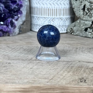 May include: A dark blue sphere, approximately 1 inch in diameter, sits on a clear plastic stand. The sphere has a speckled appearance. The background includes amethyst crystals, a white patterned pot with greenery, and a silver-colored crescent moon-shaped object.