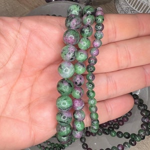 May include: Three green, pink, and black gemstone bracelets with round beads. The bracelets are stacked on top of each other on a hand.
