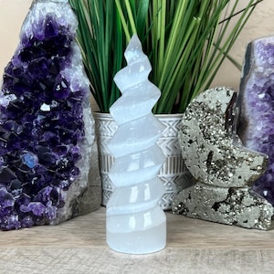 Puede incluir: Cristal de selenita blanco tallado en forma de espiral. El cristal mide aproximadamente 15 cm de alto y está sobre una superficie de madera.
