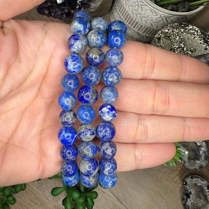 Könnte beinhalten: Ein Stapel aus drei blauen Lapislazuli-Perlenarmbändern mit silbernen Flecken. Die Armbänder bestehen aus runden Perlen und sind übereinander gestapelt.