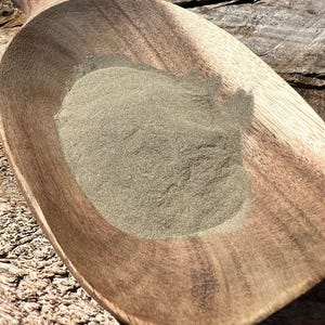Puede incluir: Una cuchara de madera llena de un montón de polvo fino de color verde claro. La cuchara es de madera marrón oscuro con un diseño liso y curvado. El polvo tiene un aspecto ligeramente texturizado.