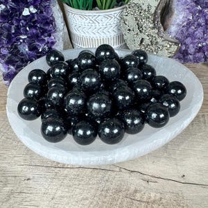 May include: A white bowl holds numerous polished black spheres with a glossy sheen. The bowl rests on a wooden surface, with amethyst crystals and other decorative items visible in the background. The spheres are approximately 2-3 cm in diameter.