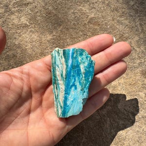 May include: A raw, unpolished mineral specimen featuring vibrant shades of turquoise and teal blue with distinct white vertical veining. The stone has a jagged, irregular shape with a textured surface, showcasing natural color variations and layers.