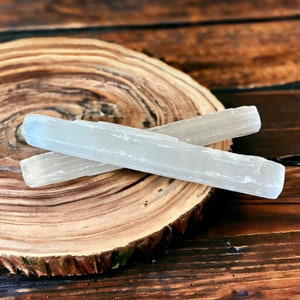 May include: Two white selenite crystal wands are displayed on a wooden slice with a natural wood grain pattern. The crystals are smooth and rectangular, with a slightly translucent appearance. The background is a dark, blurred wooden surface.