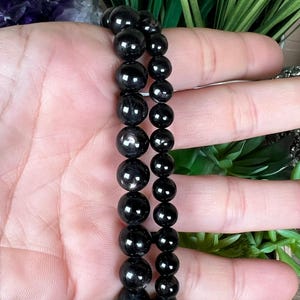 May include: Two black beaded bracelets with varying bead sizes. The beads have a glossy, reflective surface. The bracelets are displayed against a background of green foliage and a hand.