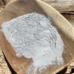Puede incluir: Una cuchara de madera llena de un polvo gris claro. La cuchara es de madera de color marrón claro y tiene un mango. El polvo parece estar finamente molido y tiene una superficie ligeramente texturizada. El fondo es una superficie de madera.