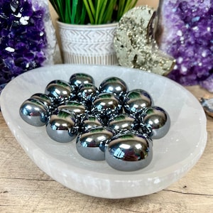 May include: A white selenite bowl filled with 15 smooth, round, dark gray hematite stones.