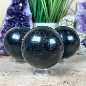 May include: Three polished black spheres on clear stands. The spheres have a reflective surface and sit on a wooden surface. The background includes amethyst geodes and green plants, suggesting a theme of natural crystals and decor.