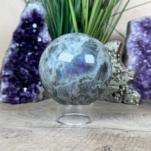 May include: A polished labradorite sphere, approximately 3 inches in diameter, displayed on a clear acrylic stand. The sphere exhibits a mix of gray, blue, and purple hues. Amethyst crystals and greenery are in the background.