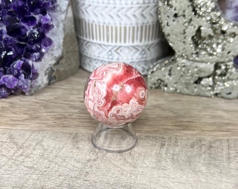 May include: A pink and white sphere with a swirling pattern, sitting on a clear stand. The sphere is made of a natural stone, possibly a type of agate.