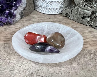 Könnte beinhalten: Eine weiße Selenit-Ladestation mit drei Kristallen: einem roten Karneol, einem klaren Quarz und einem violetten Amethyst.