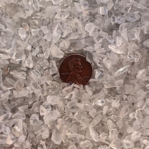 Puede incluir: Una imagen de primer plano de un montón de virutas de cuarzo transparente. Las virutas son pequeñas e irregulares en forma. Un centavo de dólar estadounidense se coloca encima de las virutas para la escala.