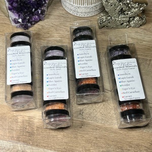 Puede incluir: Cuatro frascos de vidrio transparente que contienen minerales triturados etiquetados como "Purple Dot Alchemy Crushed Minerals" con diferentes estilos: polvo, arena, grueso y extra grueso. Cada frasco contiene una variedad de cristales de colores, incluyendo amatista, lapislázuli, apatita azul, peridoto, ojo de tigre, cornalina roja y granate.