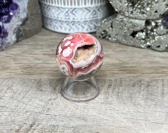 May include: A pink and white agate sphere with a natural hole in the center. The sphere is sitting on a clear stand.