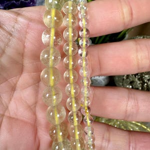 Puede incluir: Tres pulseras de cuentas con cuentas esféricas translúcidas de color amarillo. Las cuentas están ensartadas en un cordón elástico amarillo. Las pulseras varían en tamaño de cuentas. Las pulseras se sostienen en una mano.