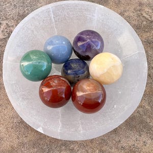 May include: Seven smooth, polished gemstone spheres in a white selenite bowl. The spheres are arranged in a circle and are in the colors of the rainbow: red, orange, yellow, green, blue, indigo, and violet.